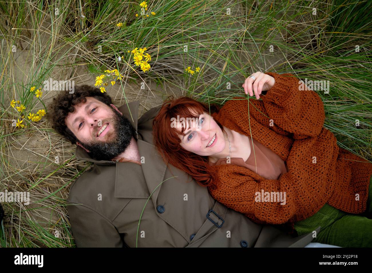 Coppia sorridente stesa sull'erba con fiori gialli intorno a loro, vestita con colori autunnali, Belgio Foto Stock