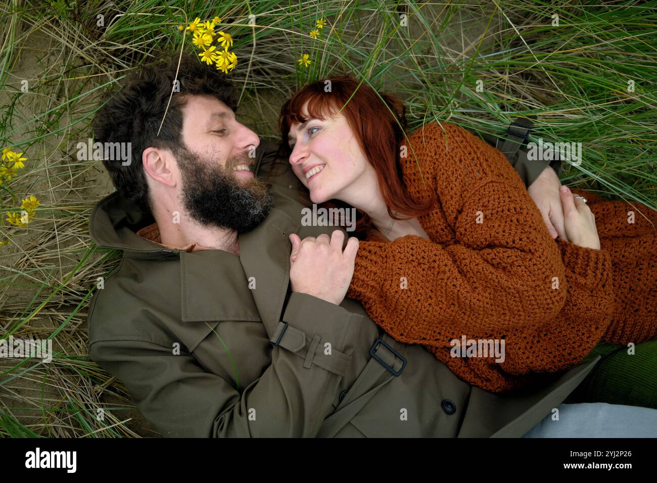 Una coppia sdraiata sull'erba, guardarsi con affetto e sorridente, circondati dal verde e dai fiori gialli. Foto Stock