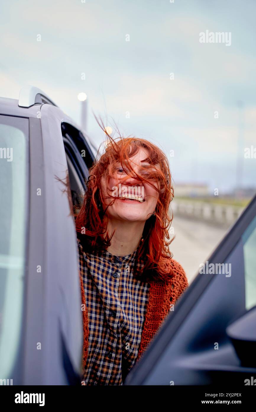 Donna rossa sorridente con una camicia a quadri e sciarpa arancione in piedi accanto a un'auto con i capelli che soffiano nel vento. Foto Stock