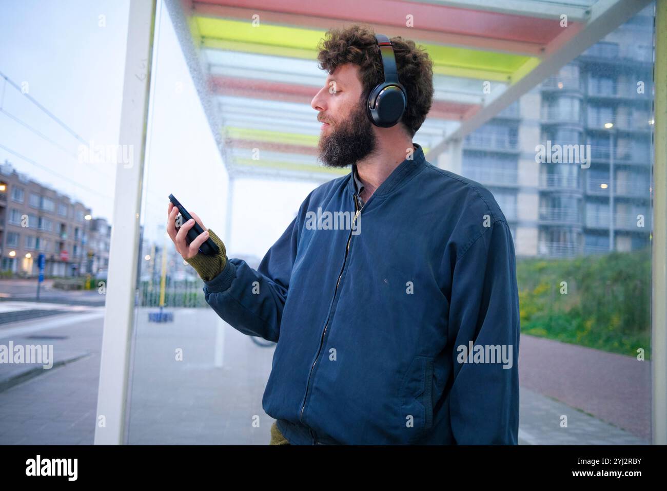 Uomo con le cuffie che usa lo smartphone alla fermata dell'autobus durante le ore al crepuscolo, Belgio Foto Stock