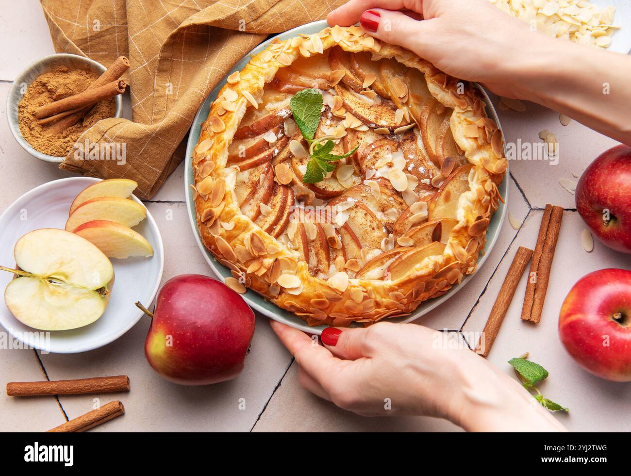 Una persona sta mettendo con cura una galette di mele appena sfornata su un tavolo circondato da mele affettate, bastoncini di cannella e fiocchi di mandorle. Il tiepido kitch Foto Stock