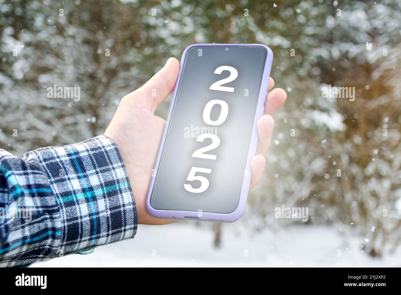 Una mano che tiene in mano uno smartphone che visualizza il numero 2025 sullo schermo, simboleggiando il nuovo anno. Lo sfondo presenta una scena invernale Foto Stock