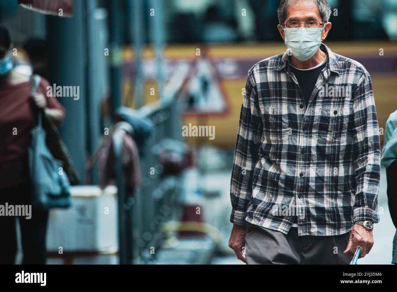 Un uomo più anziano con una camicia a quadri e una maschera facciale passa davanti alla gente in una vivace strada di Hong Kong, circondato da paesaggi urbani alla luce del pomeriggio. Foto Stock