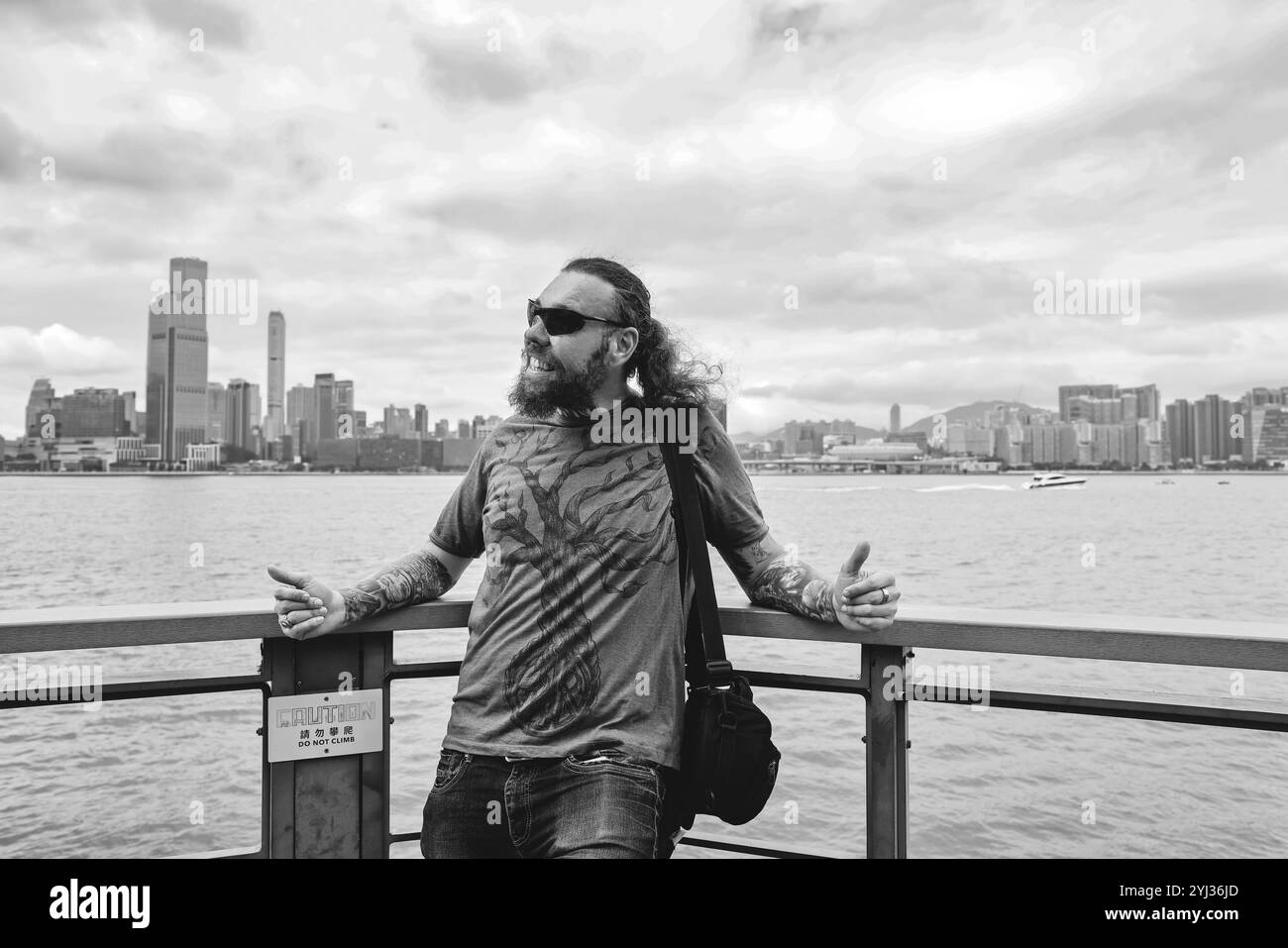 Un turista si rilassa sul lungomare di Hong Kong, mostrando l'impressionante skyline della città sotto un cielo coperto. Foto Stock