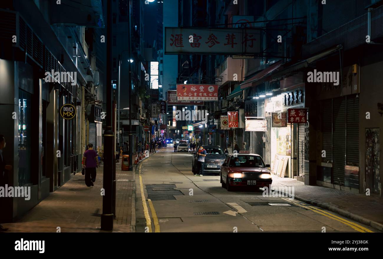 Un taxi attraversa una strada poco illuminata di Hong Kong, circondata da negozi chiusi e cartelli illuminati in una notte movimentata. Foto Stock