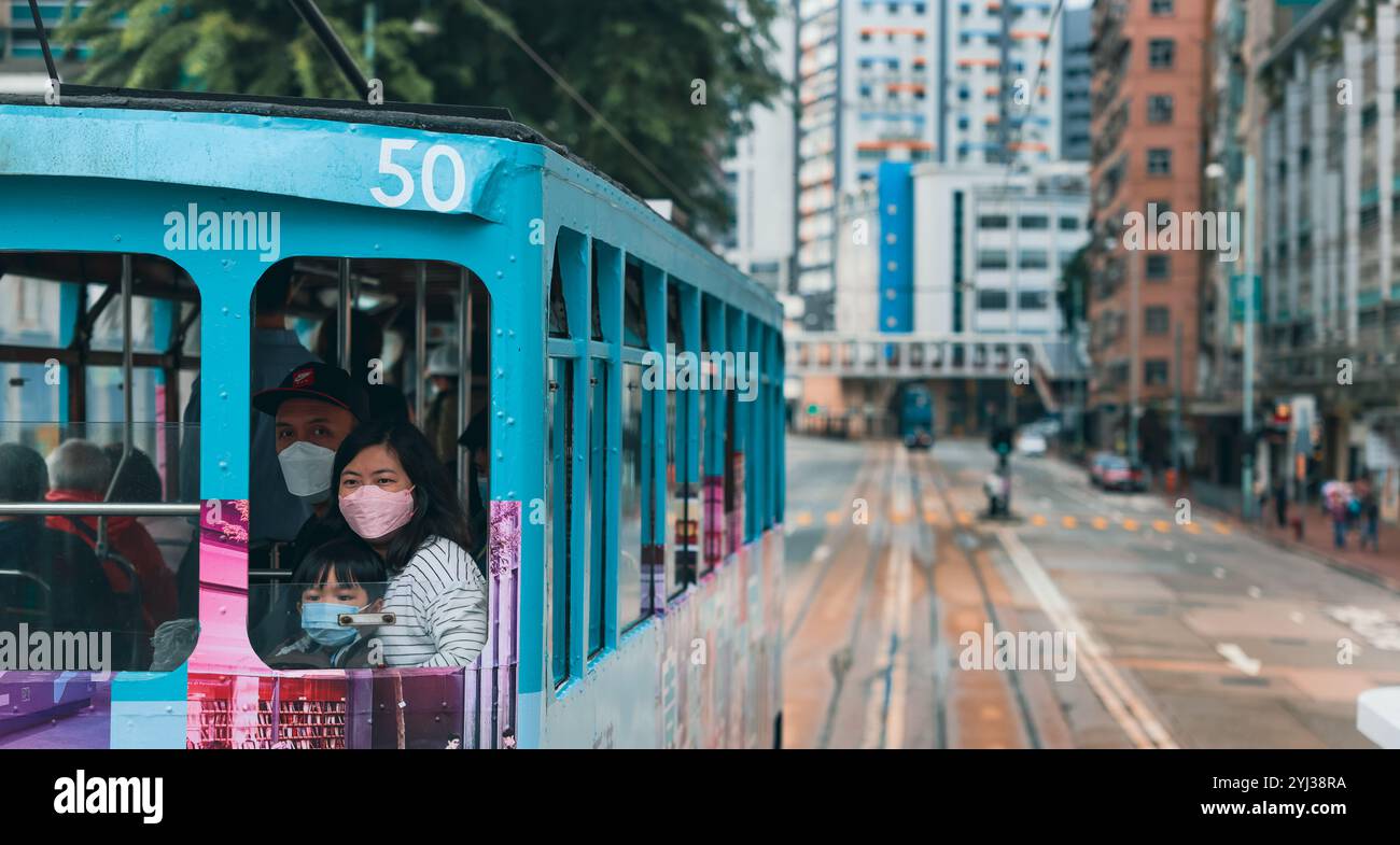 Un vivace tram viaggia attraverso le strade di Hong Kong, con i passeggeri che indossano maschere e si godono il paesaggio urbano. Foto Stock