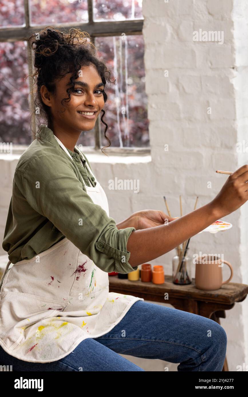 Donna sorridente che dipinge a casa, tenendo pennello e tavolozza, godendo della creatività Foto Stock