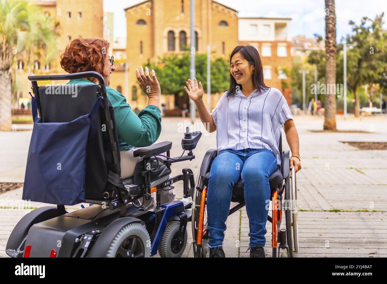 Due donne multietniche con disabilità in sedia a rotelle che salutano per strada Foto Stock