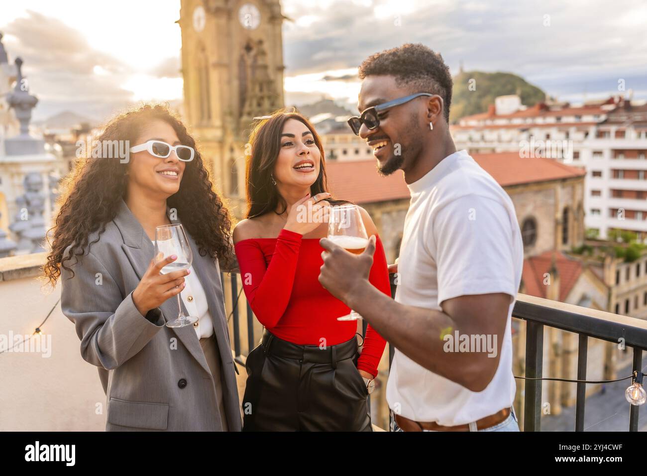Amici multietnici che chiacchierano e bevono vino sul tetto durante il tramonto con vista sulla città Foto Stock