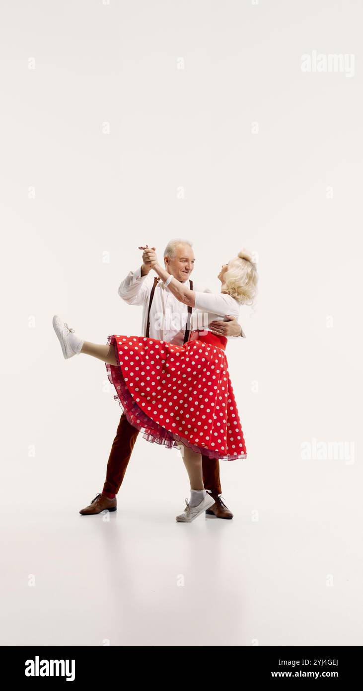 Un uomo e una donna senior si impegnano in una danza vivace, vestiti con abiti d'ispirazione vintage isolati su sfondo bianco Foto Stock