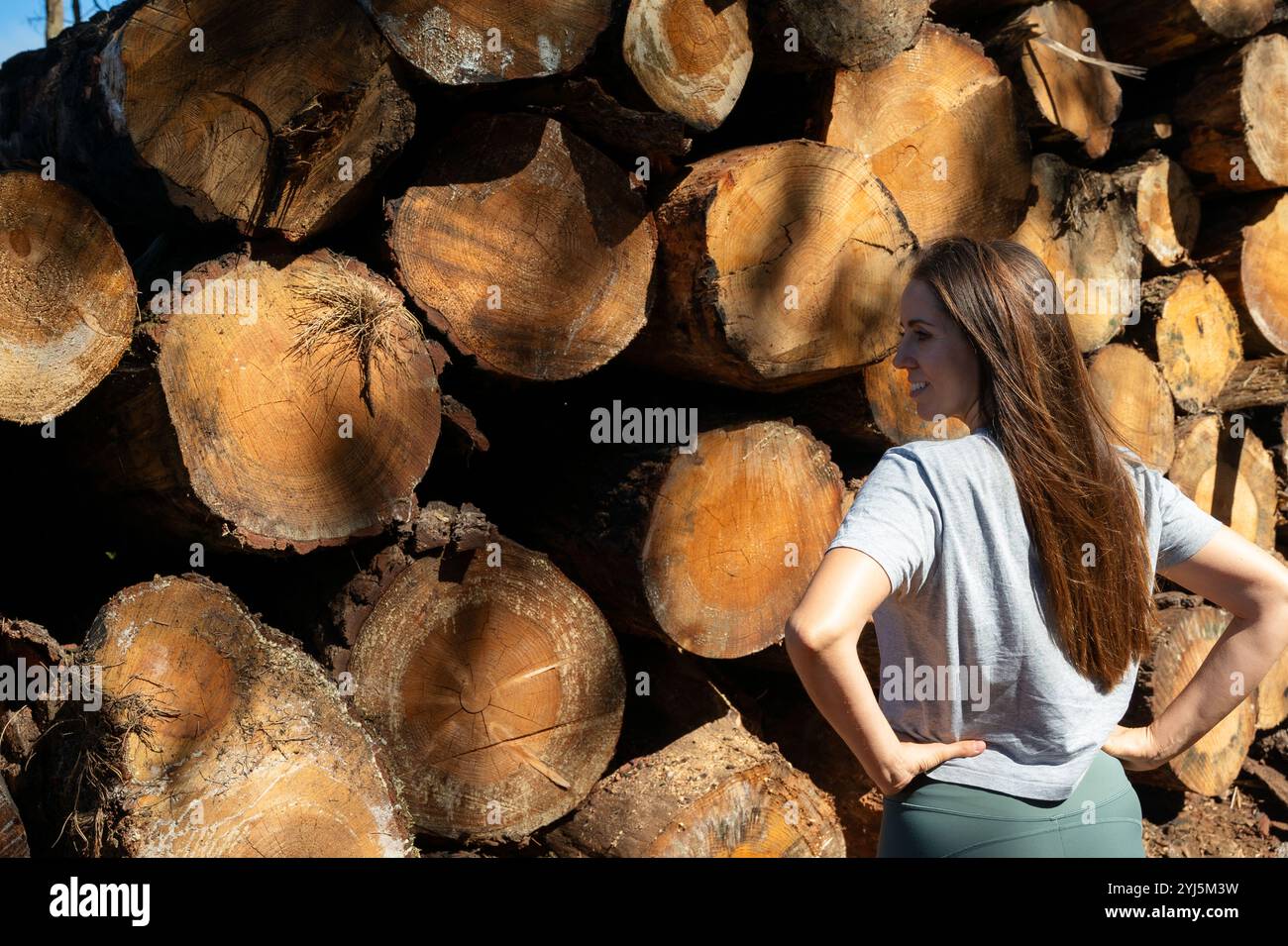 Una donna sta di fronte a una pila di tronchi, guardando la telecamera. La scena ha un aspetto rustico e naturale, con i tronchi disposti in modo da suggellare Foto Stock