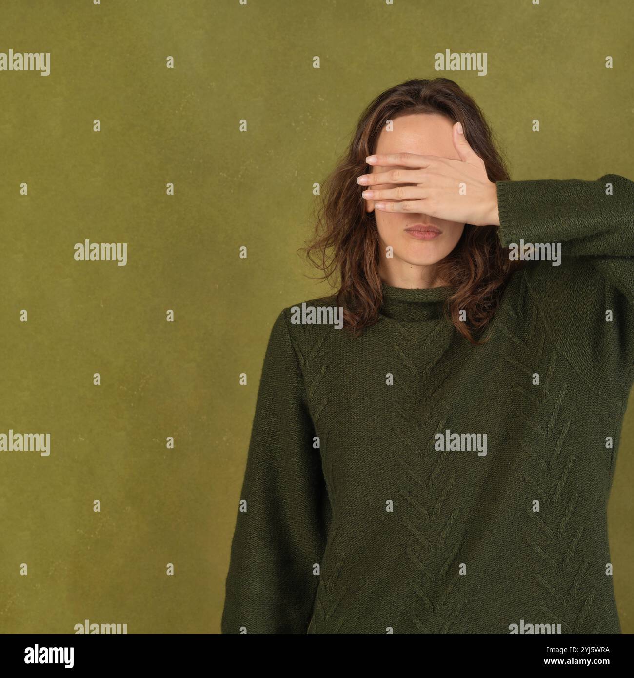 Una donna con un maglione verde copre gli occhi su uno sfondo di oliva Foto Stock