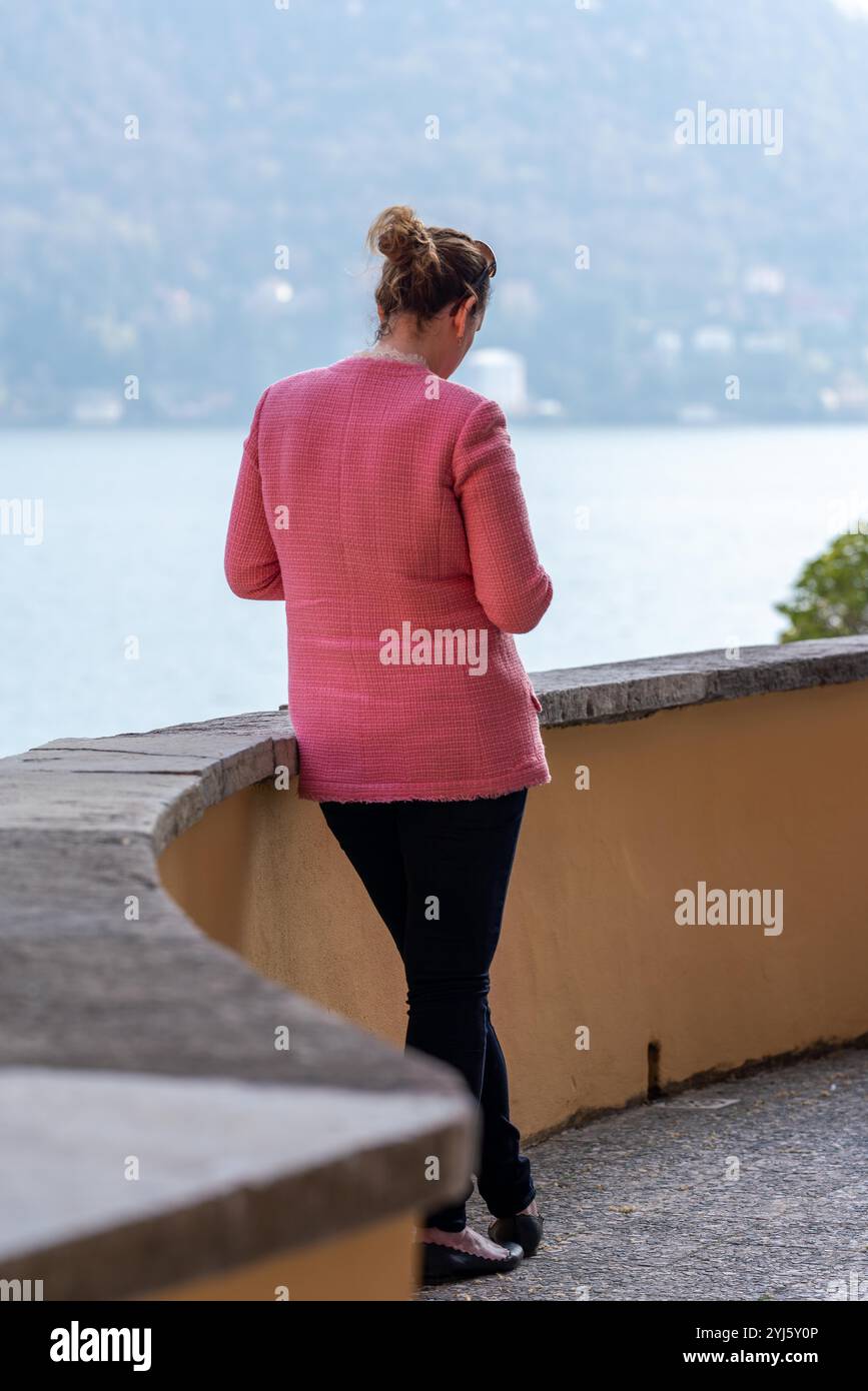 La signora con la giacca rosa rivolta verso l'esterno studia il suo telefono sulla veranda dell'hotel Villa d'Este che si affaccia sul lago di Como. Foto Stock