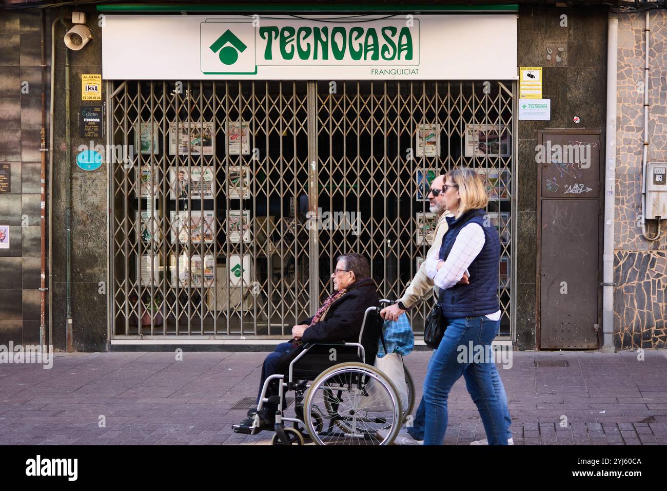 Montcada i Reixac. Spagna - 13 novembre 2024: Un uomo spinge una persona su una sedia a rotelle oltre un ufficio immobiliare Tecnocasa chiuso con cancello metallico. La S Foto Stock