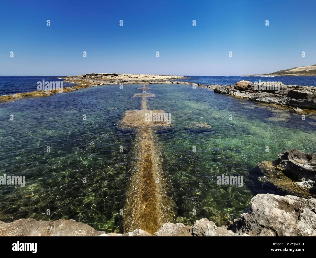 Piscina di roccia naturale limpida con acqua verde-blu lungo una costa rocciosa sotto un cielo luminoso, Malta. Foto Stock