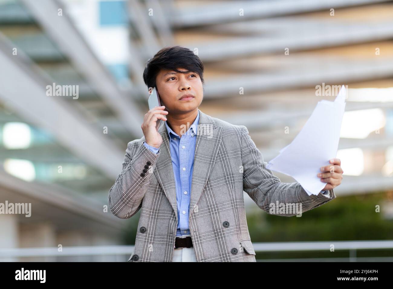 Un giovane asiatico si trova all'aperto, indossando un blazer grigio e una camicia blu, impegnato in una conversazione telefonica mentre tiene in mano documenti importanti, mostrando un Foto Stock