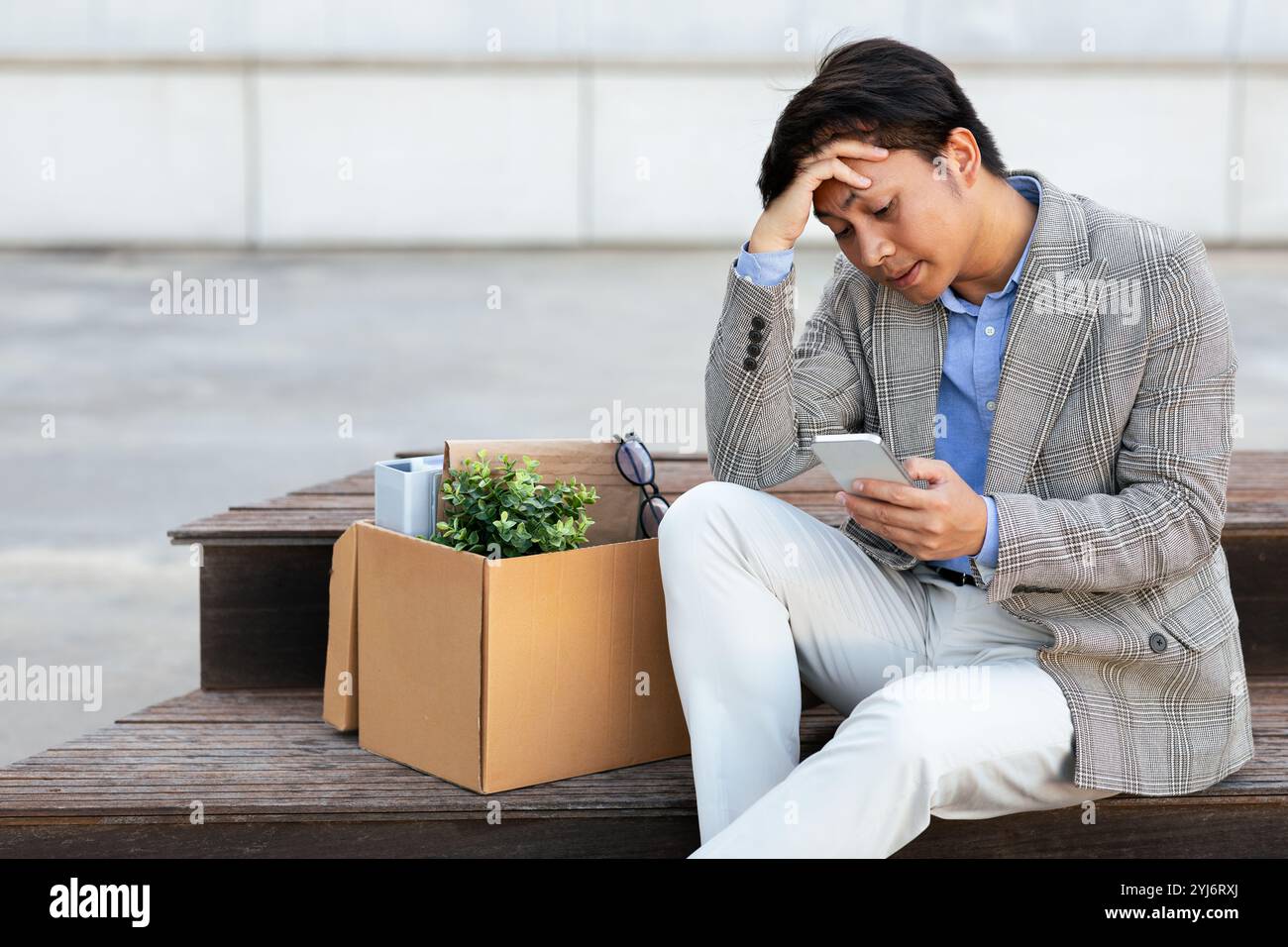 Un uomo asiatico senza lavoro con un blazer grigio e pantaloni bianchi siede su una panchina di legno. Ha i suoi effetti personali in una scatola di cartone accanto a lui. Guarda in basso la sua Foto Stock