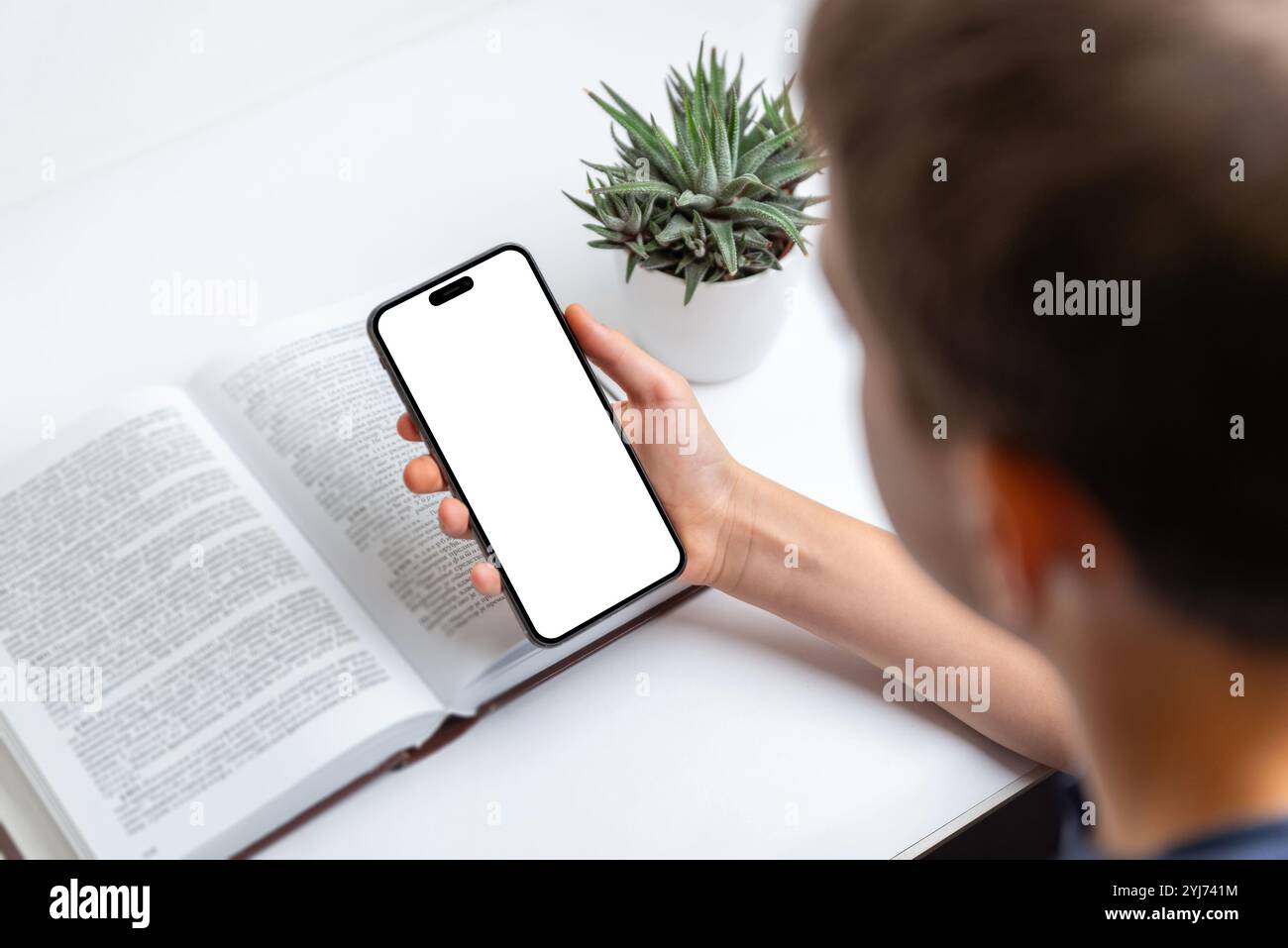 Bambino che tiene il telefono accanto a un libro, schermo isolato, ideale per i mockup e le presentazioni di app. Perfetto per la tecnologia, l'apprendimento, l'istruzione e il digitale Foto Stock