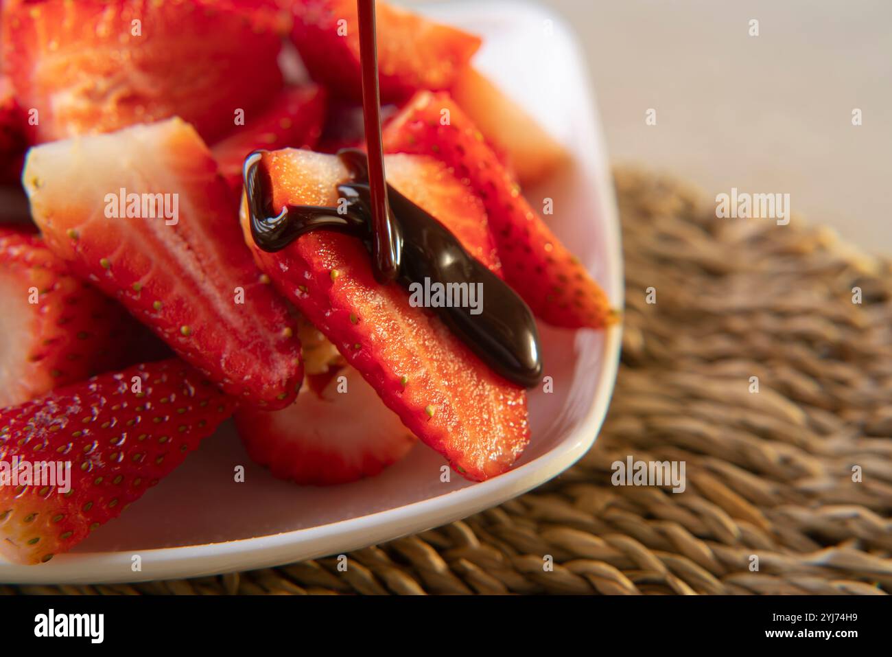 Primo piano di cioccolato che gocciola su una fragola su un piatto quadrato bianco con fragole tritate sul tavolo di vimini Foto Stock
