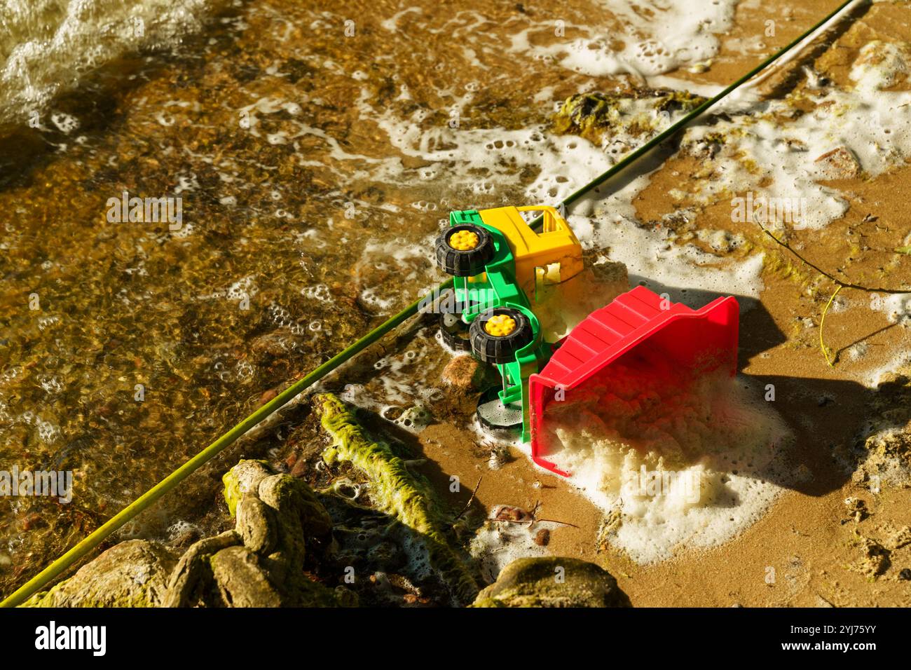 Un camion giocattolo giallo brillante e verde poggia su una spiaggia sabbiosa, parzialmente sommersa da acqua schiumosa, creando un'atmosfera invitante e giocosa sotto Foto Stock