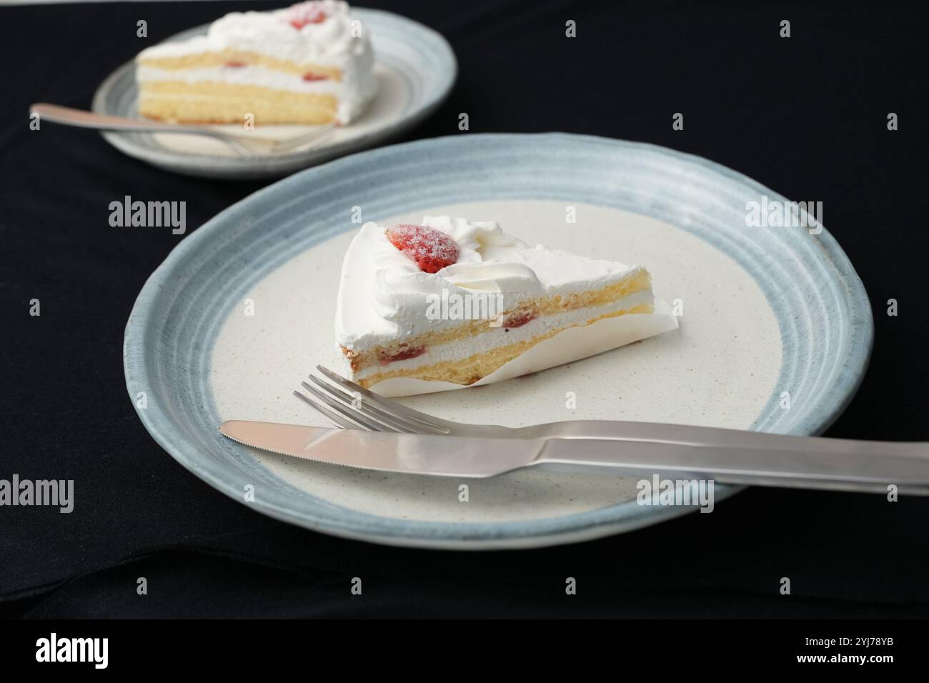 Piccola torta di fragole giapponese, forchetta e coltello ciascuno su due eleganti piatti grandi e piccoli in stile giapponese su un panno nero su un tavolo bianco. Foto Stock