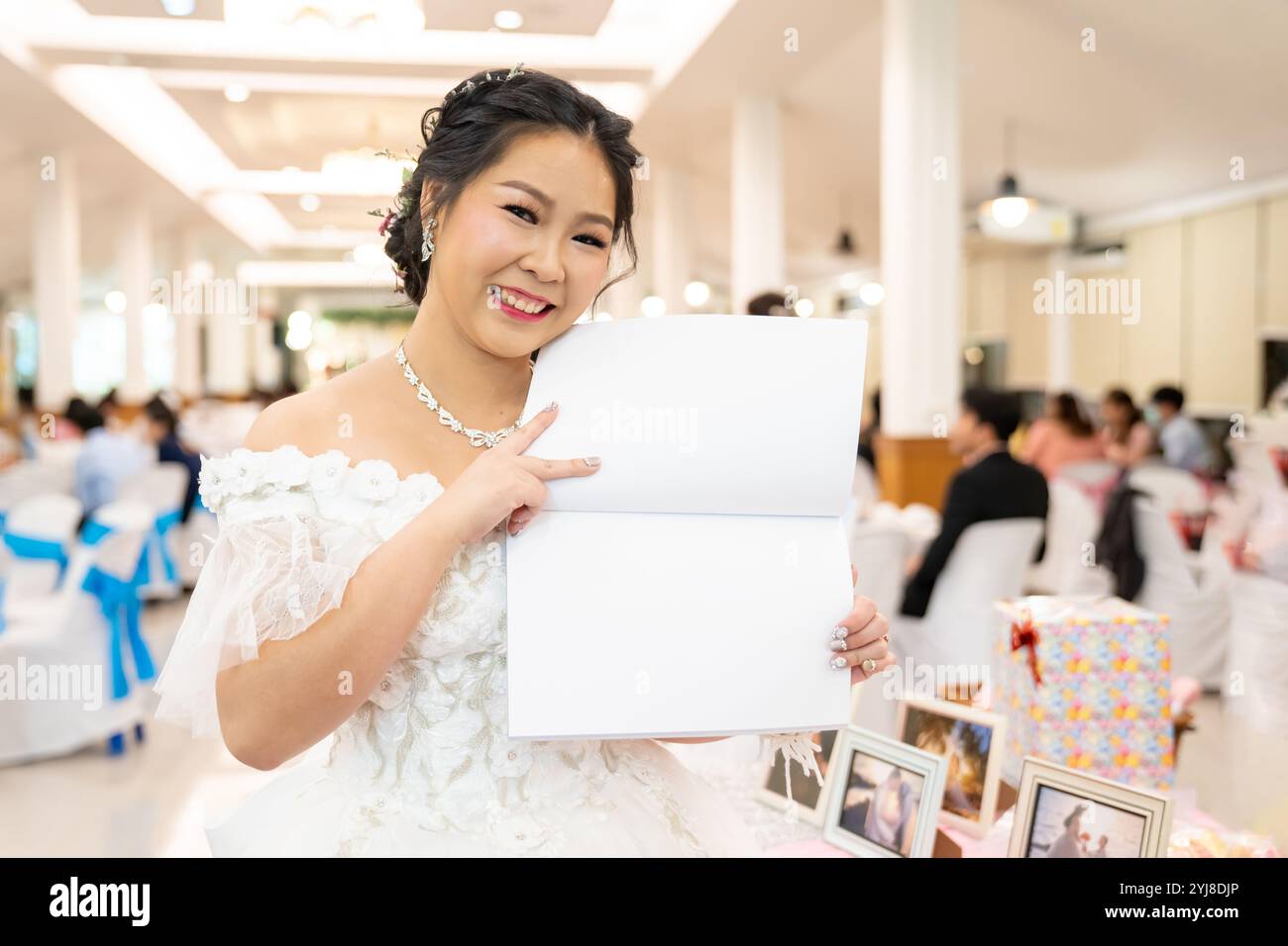 Un ritratto di una ragazza felice in un vestito bianco che tiene aperto un libro vuoto vicino ad un tavolo di ricevimento in un evento, organizzato in un'elegante sala d'albergo, con un Foto Stock