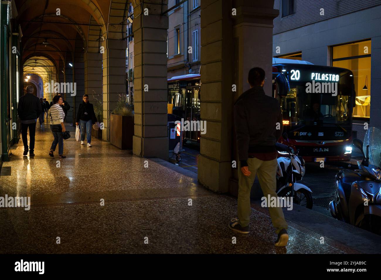Bologna, Italia. 8 ottobre 2024 - persone che camminano sotto archi storici con un autobus in strada la sera Foto Stock