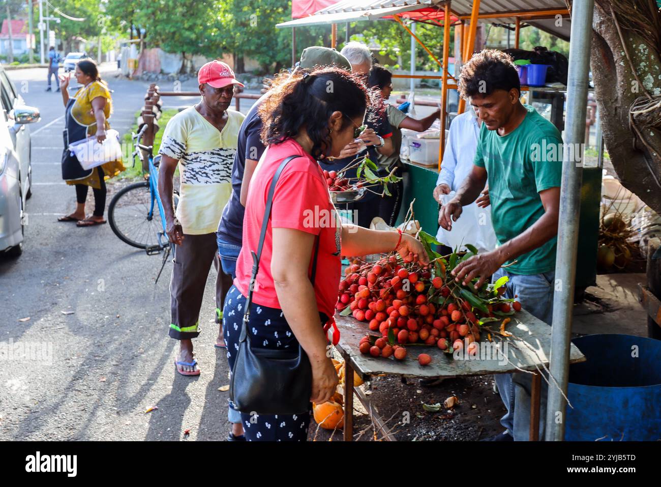 Un gruppo eterogeneo di individui si riunisce intorno a un banco di frutta a Mauritius, esaminando e acquistando vari litchi e altri frutti. Foto Stock