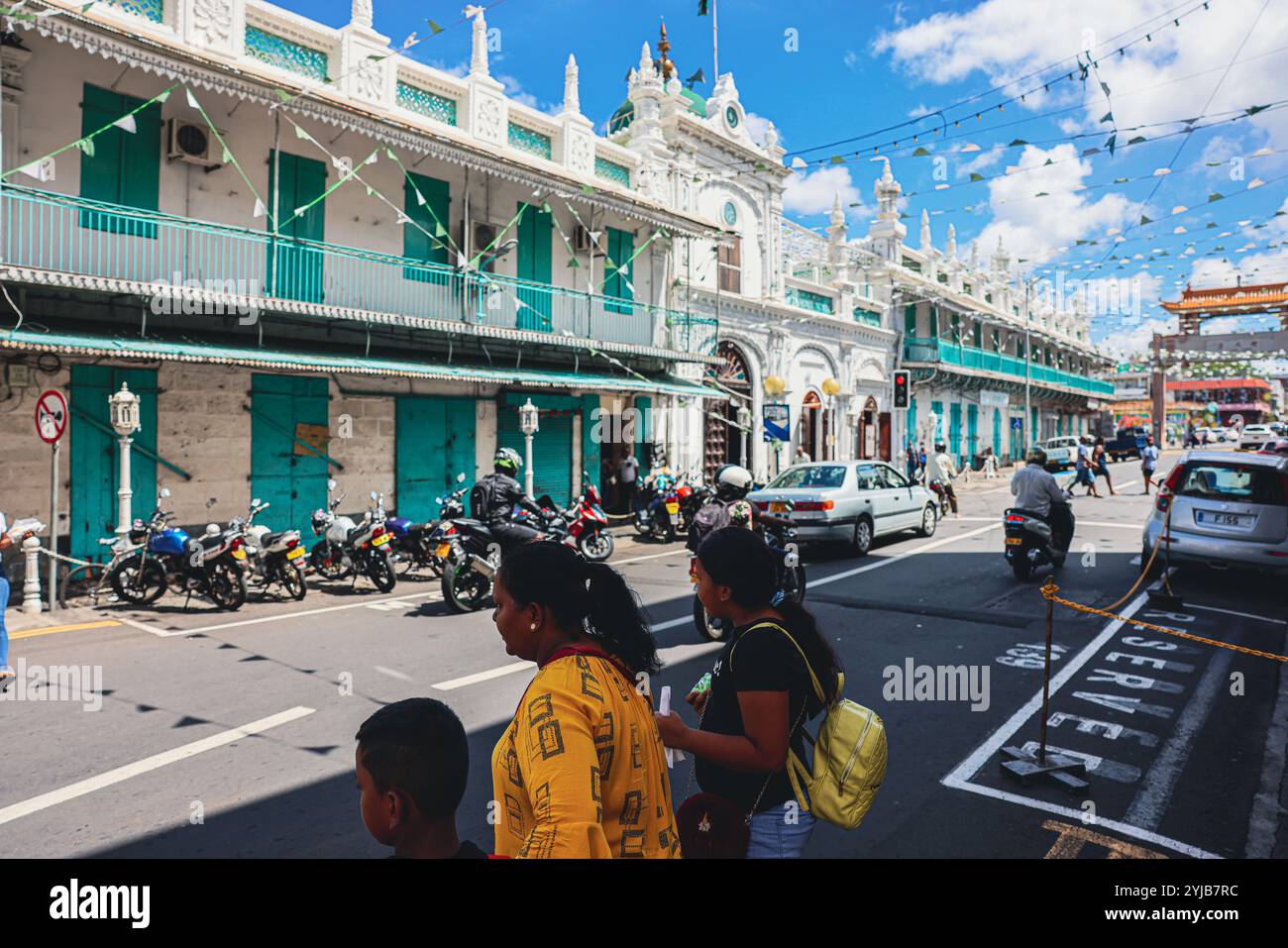 Un gruppo variegato di individui in piedi nella strada di Port Louis City con vari edifici e una moschea sullo sfondo. Foto Stock