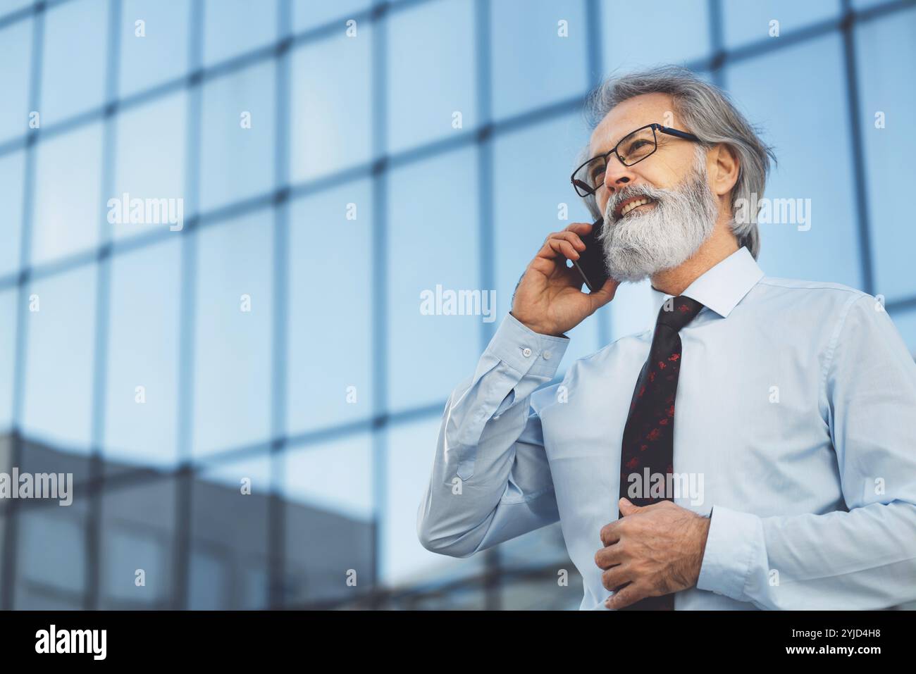 Uomo d'affari anziano con capelli grigi e barba che parla al telefono fuori in una giornata di sole. CEO alla chiamata fuori Foto Stock