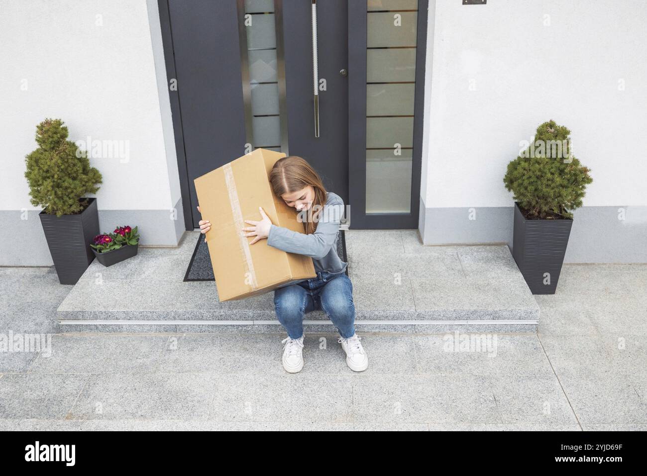 Eccitante donna caucasica seduta davanti alla porta d'ingresso con una grande scatola di cartone appena arrivata per posta. Donna che riceve un pacchetto eccitante Foto Stock