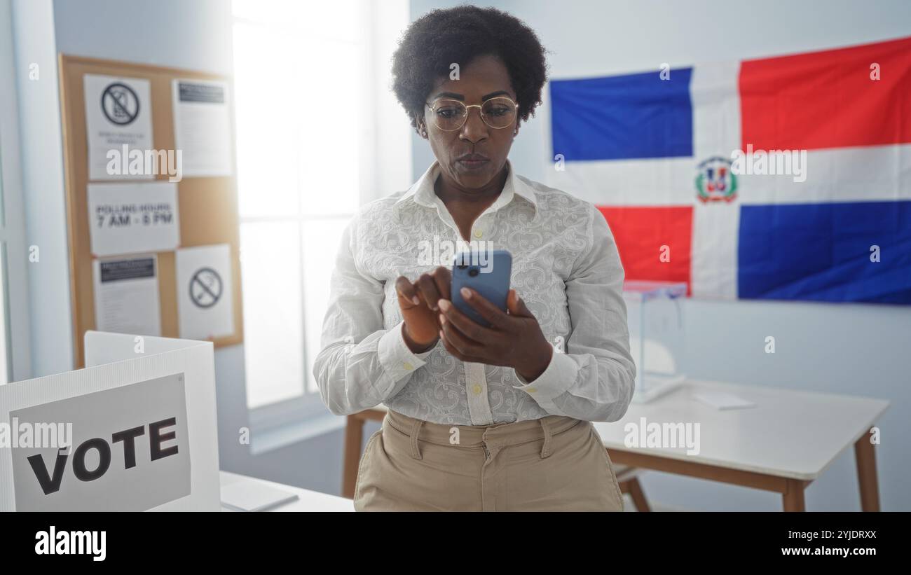 Donna afroamericana in una sala del college elettorale nella repubblica dominicana che usa il suo telefono con un segno di voto e una bandiera nazionale sullo sfondo. Foto Stock