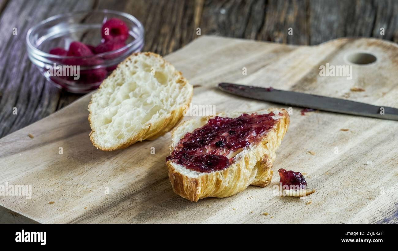 Un pezzo di croisant con marmellata e un coltello su un tagliere di legno. Sullo sfondo è presente una piccola ciotola di lamponi. Foto Stock