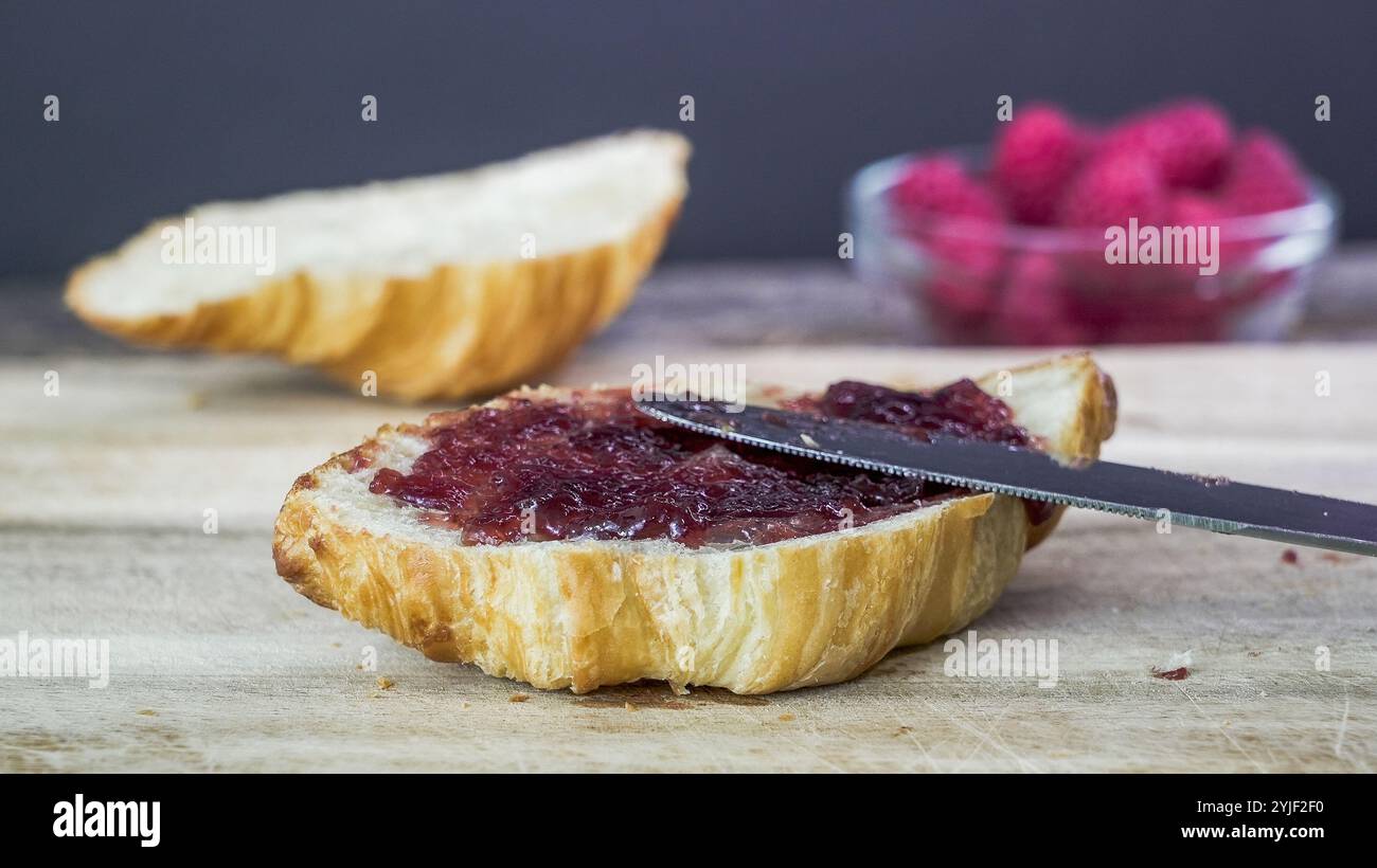 Un coltello distribuisce una briciola di marmellata rossa su un croissant. Un altro pezzo di pane poggia sul tagliere nelle vicinanze. Foto Stock