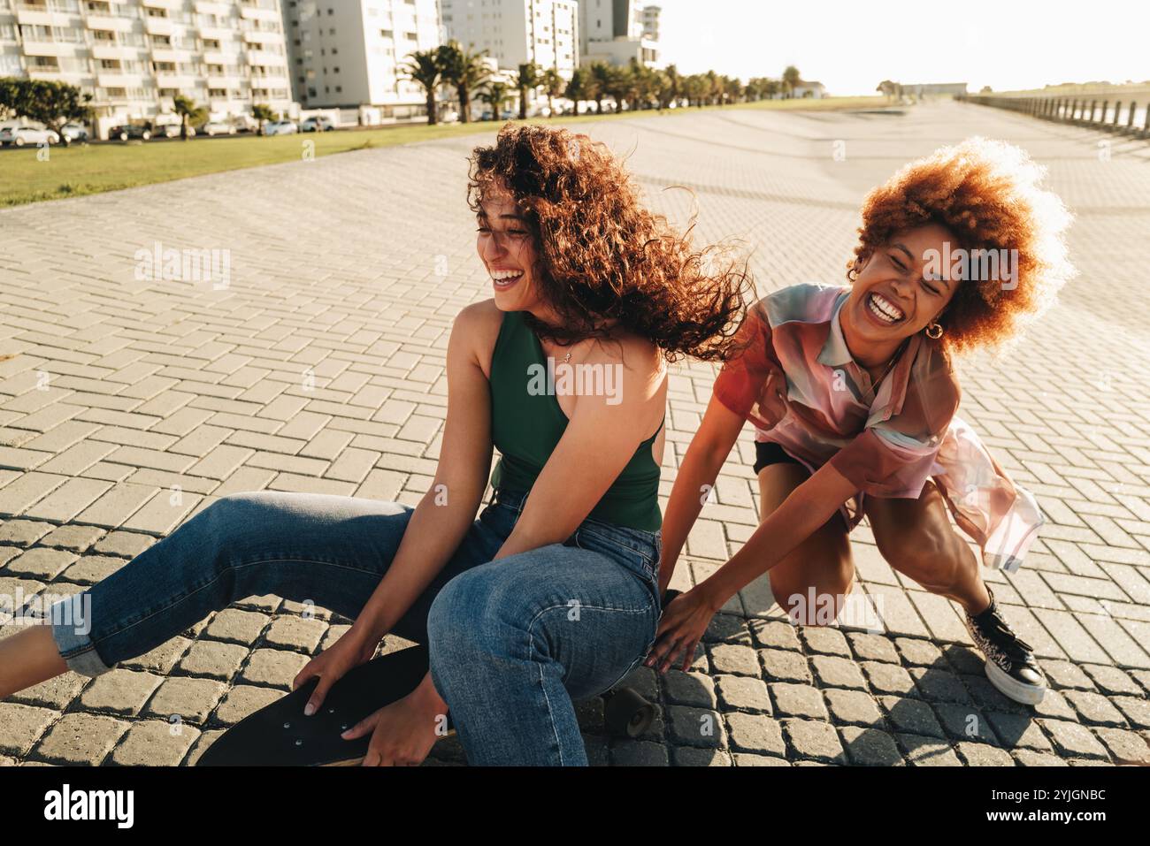 Due amici ridono e fanno uno skateboard insieme in una giornata di sole, godendosi divertimento e amicizia in un ambiente urbano. Foto Stock