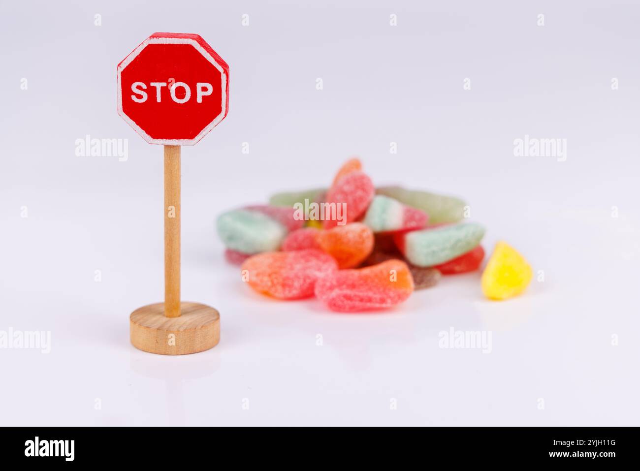 Un segnale di stop in legno posto accanto a una pila di caramelle gommose di diversi gusti, su sfondo bianco, creando una scena giocosa e colorata Foto Stock