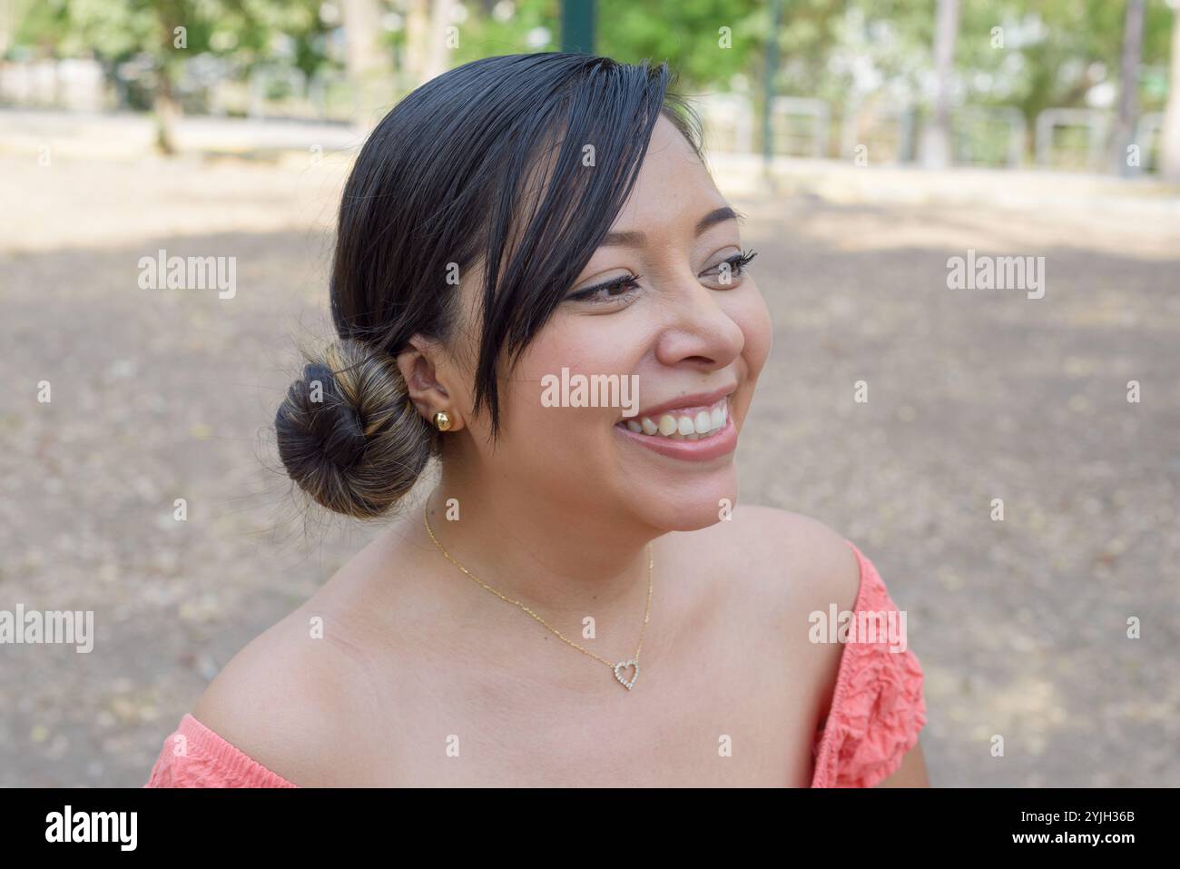 Una giovane donna con i capelli tirata indietro in un panino sorride brillantemente, indossando un top rosa a spalla. Foto Stock
