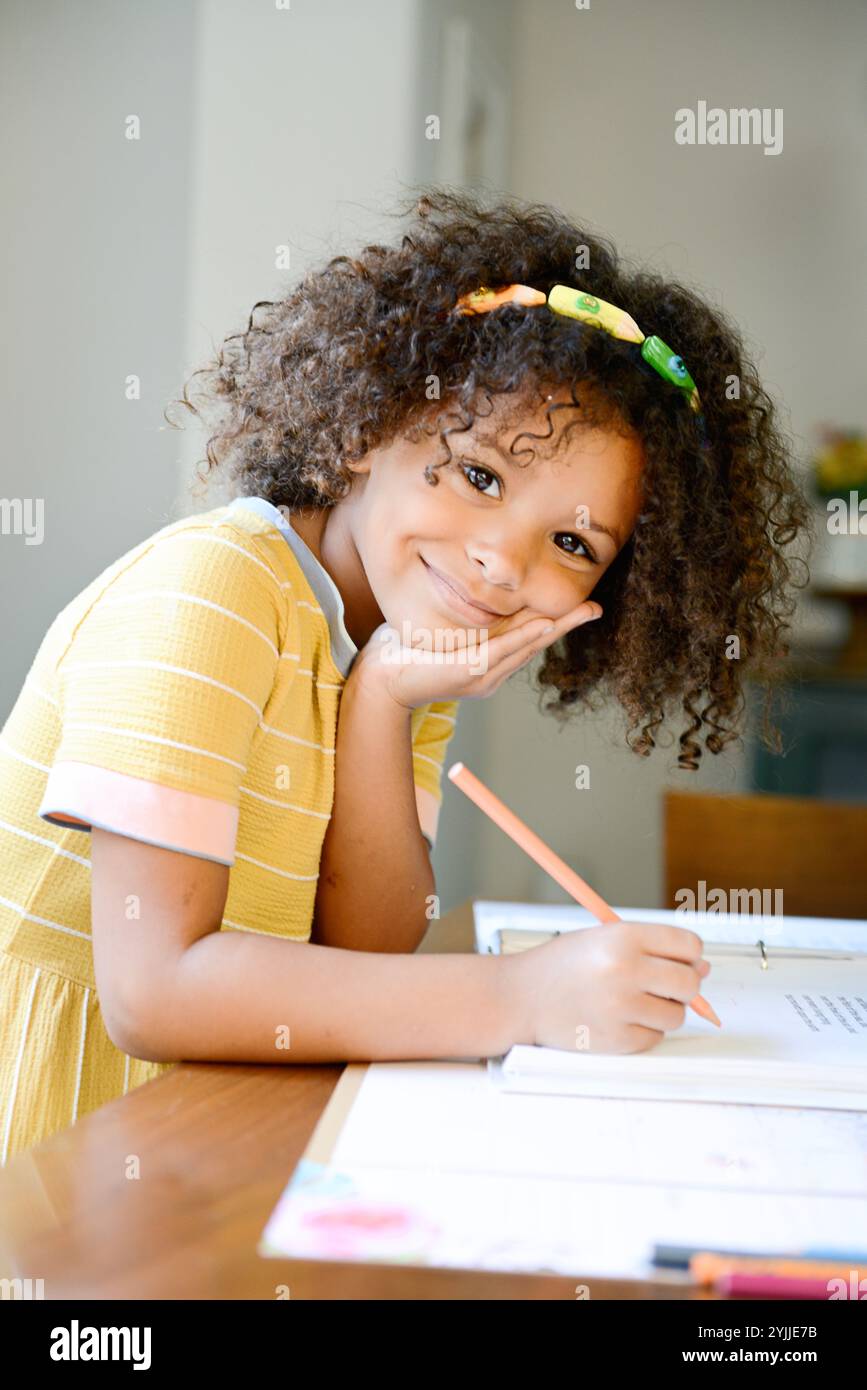 Bambino sorridente in abito giallo che scrive con una matita a un tavolo Foto Stock