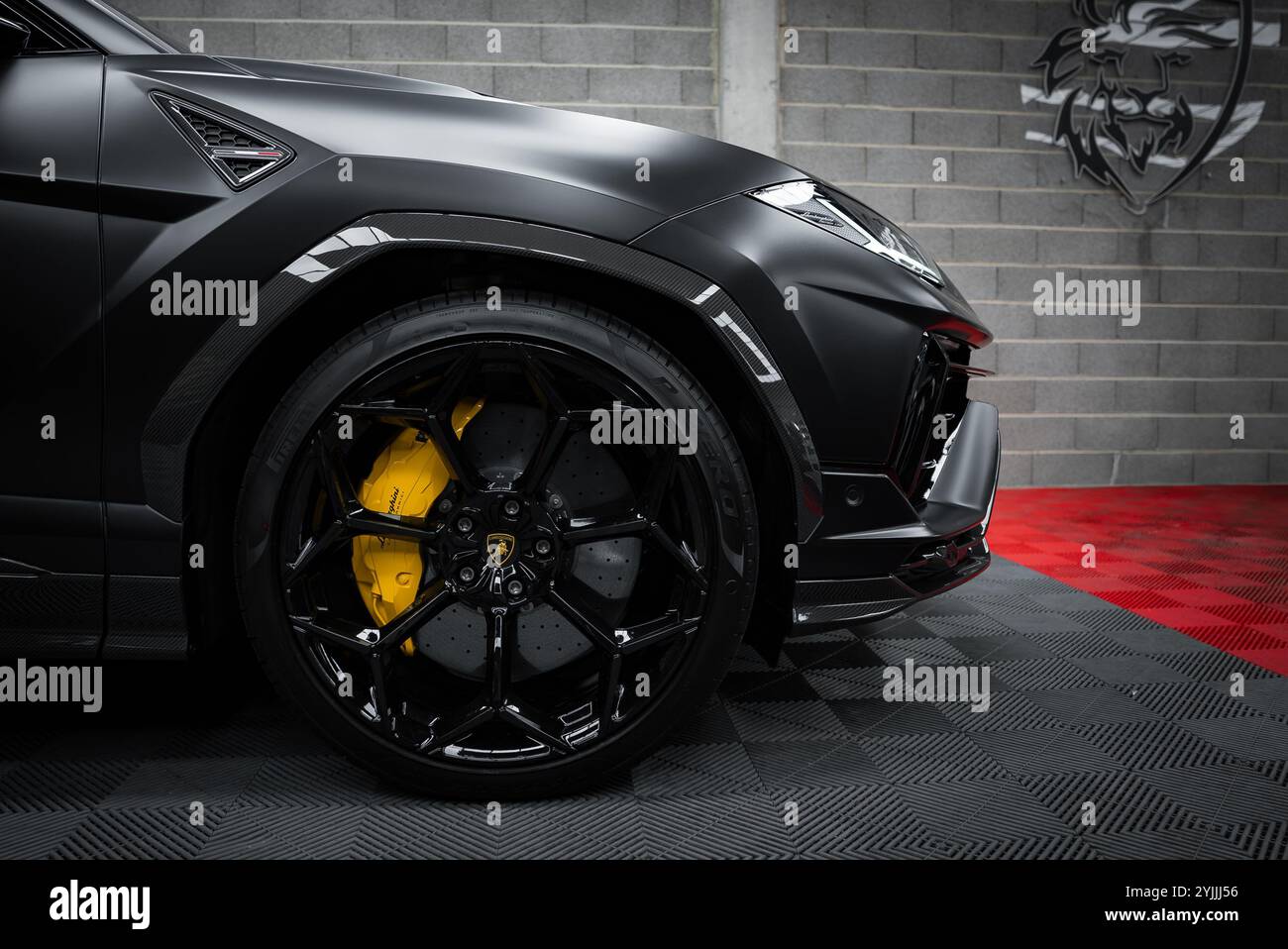 L'immagine mostra un primo piano della ruota anteriore di una Lamborghini Urus Performante con cerchioni in lega nera e pinze dei freni gialle, appoggiate su un mattone grigio W. Foto Stock