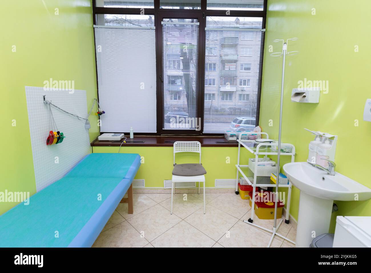 Una luminosa sala visite mediche con parete d'accento verde, letto paziente blu, finestra con persiane, sedia, forniture mediche su un carrello, stativo IV e peccato Foto Stock