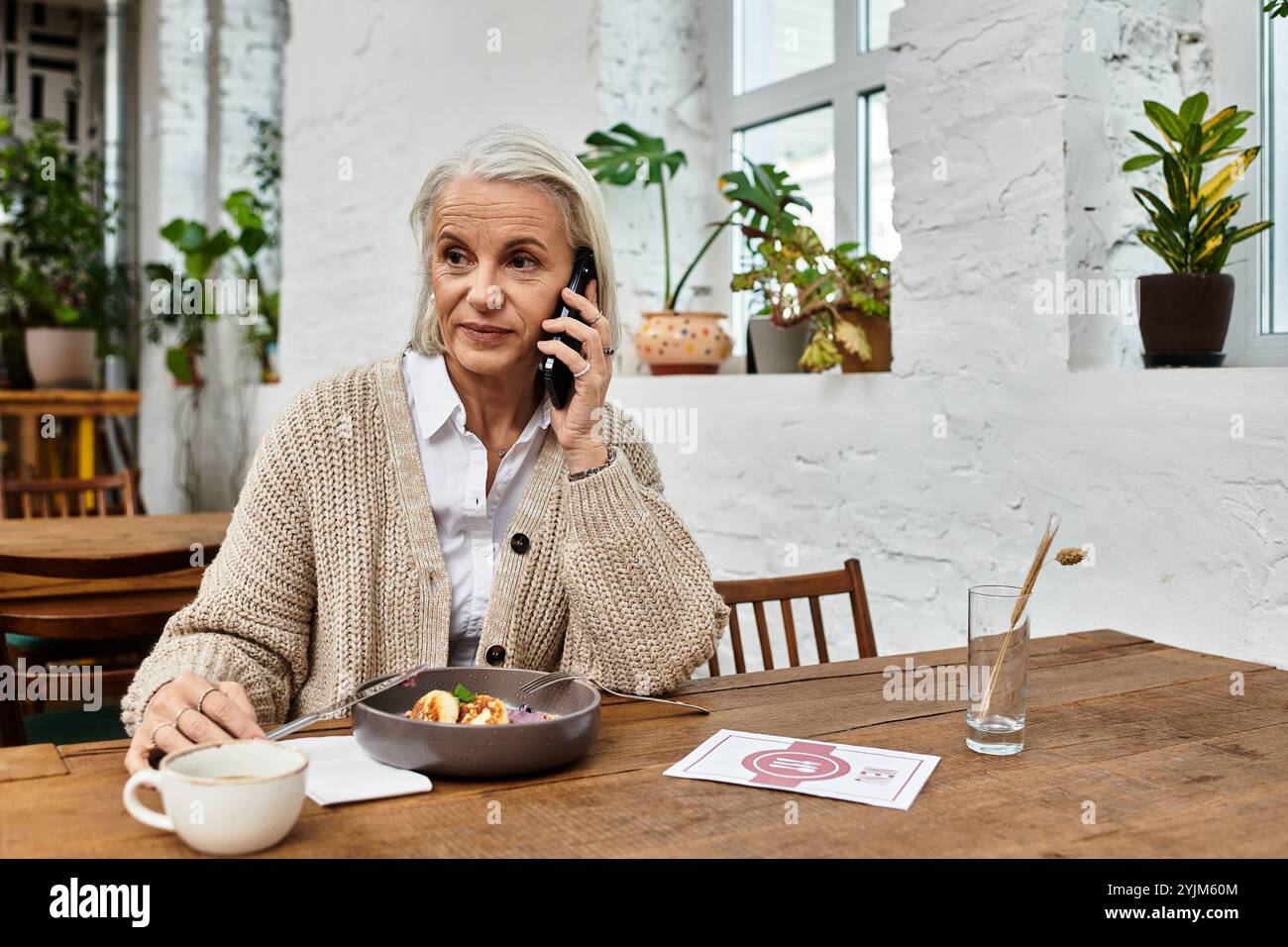 Una bella donna matura si impegna in una conversazione telefonica ponderata, assaporando un pasto delizioso. Foto Stock