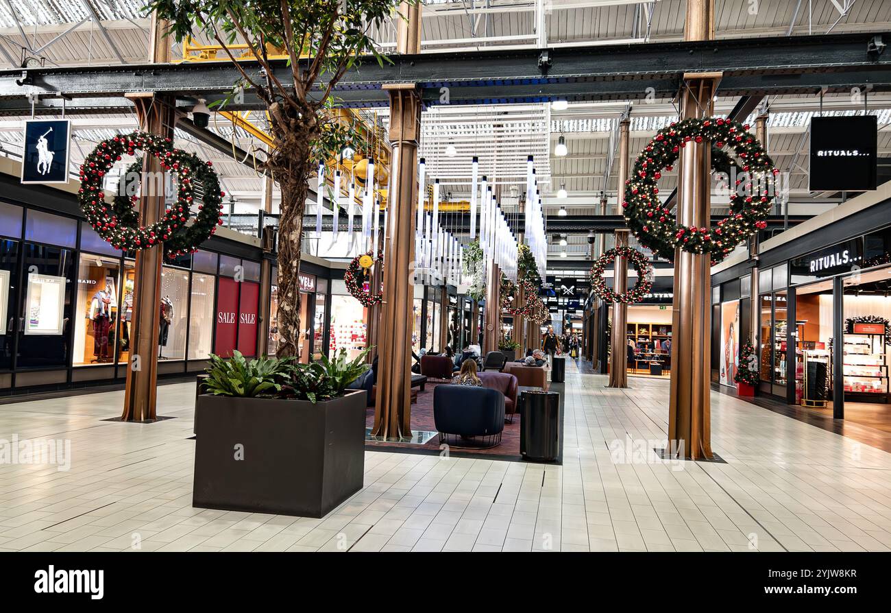 La corona natalizia decora il corridoio di un centro commerciale mentre la gente passeggia accanto ai negozi durante le festività natalizie del centro outlet Swindon 2024 Foto Stock