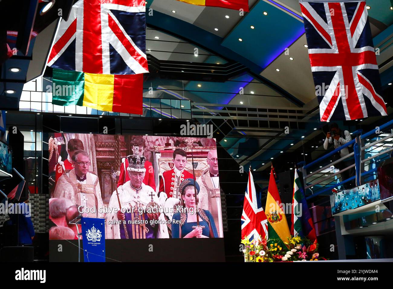 El alto, BOLIVIA; 15 novembre 2024: Immagini dell'incoronazione di re Carlo III sono esposte su un grande schermo mentre l'inno nazionale viene suonato all'inizio di un evento organizzato dall'Ambasciata britannica in Bolivia per commemorare il compleanno di re Carlo III (che era ieri). L'evento è stato anche per promuovere le relazioni culturali e il commercio tra la Bolivia e il Regno Unito. Le bandiere tricolori boliviane e le jacks unioniste pendono dal soffitto della sala eventi. Foto Stock