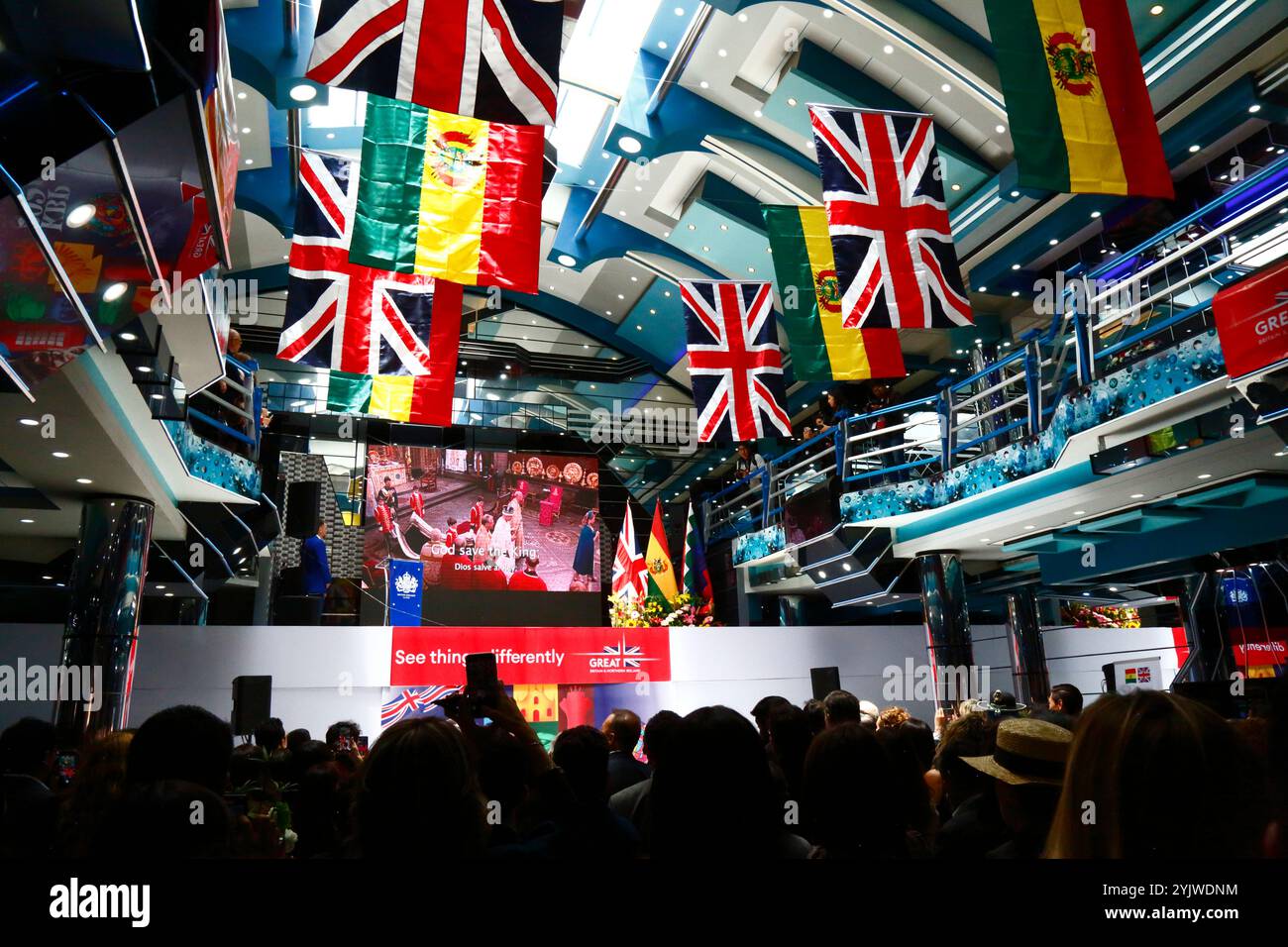 El alto, BOLIVIA; 15 novembre 2024: Immagini dell'incoronazione di re Carlo III sono esposte su un grande schermo mentre l'inno nazionale viene suonato all'inizio di un evento organizzato dall'Ambasciata britannica in Bolivia per commemorare il compleanno di re Carlo III (che era ieri). L'evento è stato anche per promuovere le relazioni culturali e il commercio tra la Bolivia e il Regno Unito. Le bandiere tricolori boliviane e le jacks unioniste pendono dal soffitto della sala eventi. Foto Stock