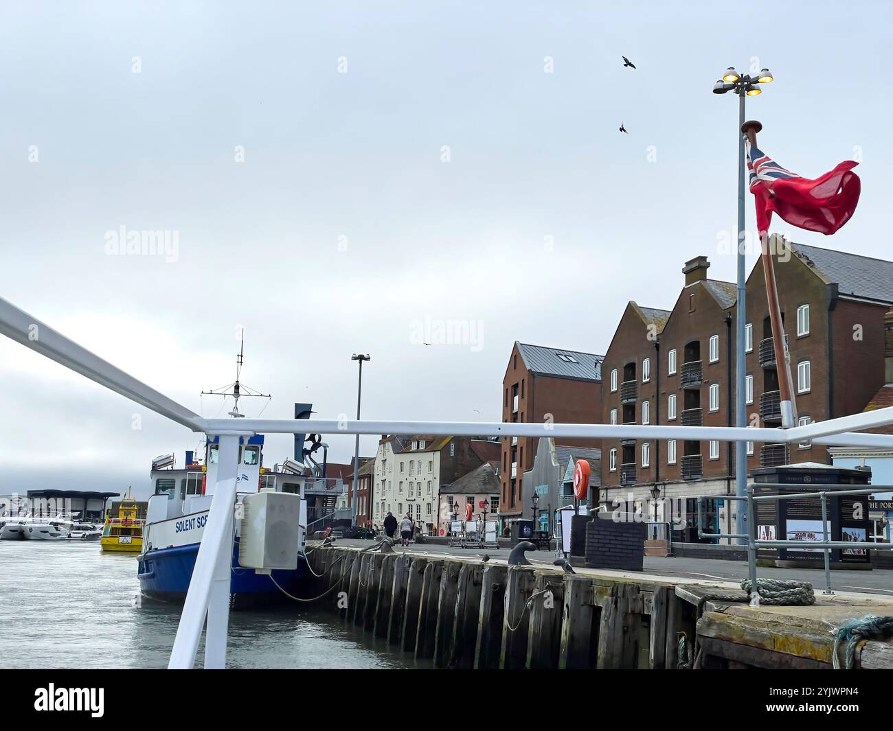 Poole, Regno Unito, 11 aprile 2024. le barche attraccavano accanto a edifici storici in mattoni, con una bandiera vibrante che sventolava nella brezza. La tranquilla posizione del porto Foto Stock