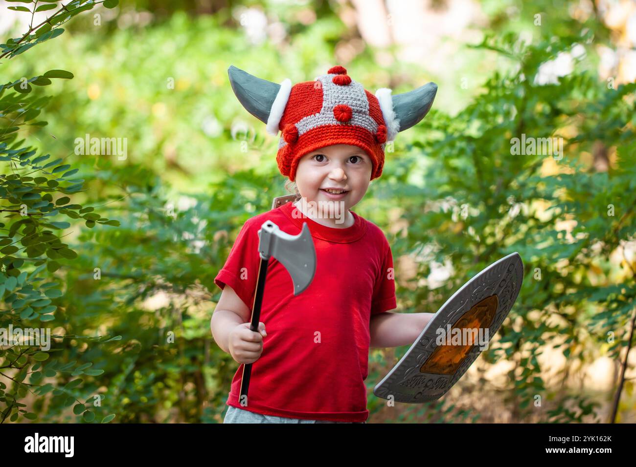 Ragazzo carino che gioca con la spada e lo scudo fuori. Idee per giochi di ruolo per bambini. Cappello vichingo fai da te. Stile di vita Foto Stock