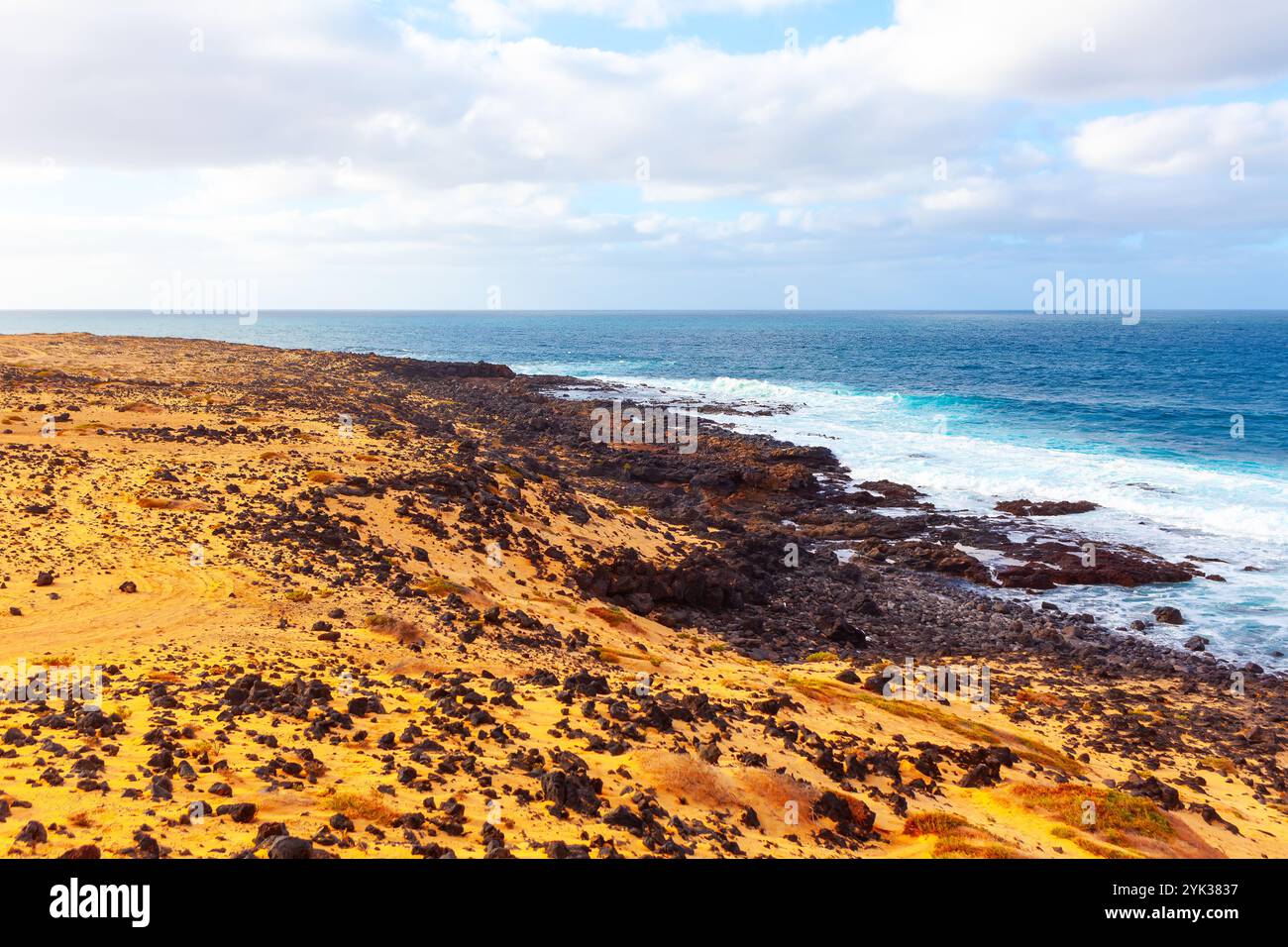 Paesaggio costiero nelle Isole Canarie con una costa rocciosa, terreno sabbioso e arido e onde oceaniche che si infrangono contro rocce scure. Il sk parzialmente nuvoloso Foto Stock