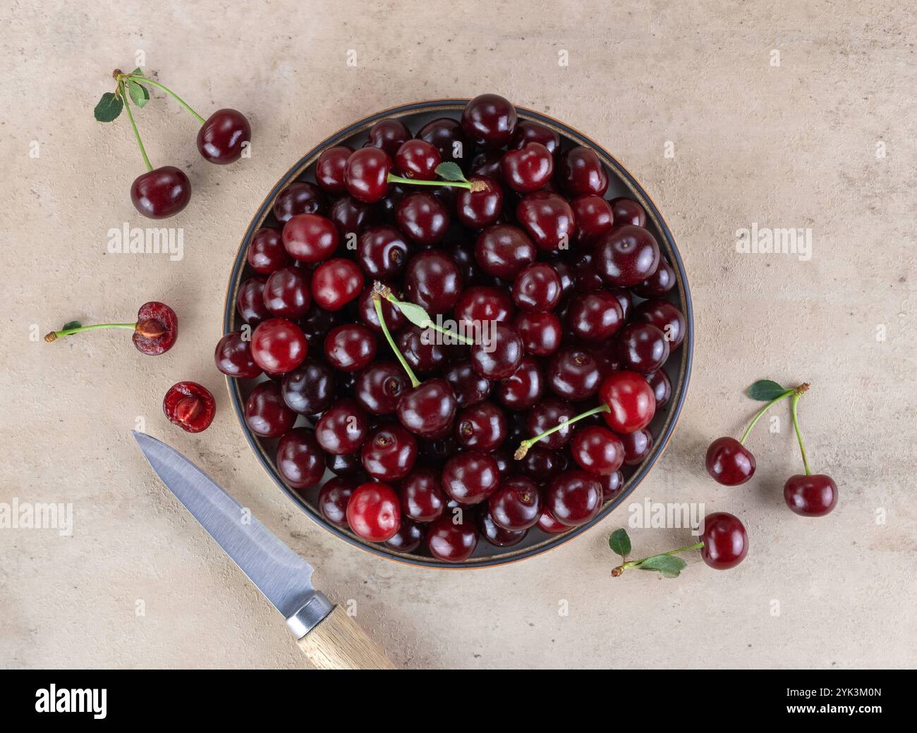 Ciotola di ciliegie rosse fresche e coltello su sfondo beige. Vista dall'alto Foto Stock