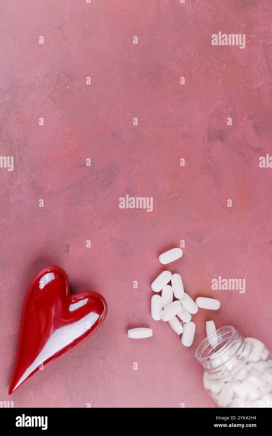 Cuori rossi e bianchi in porcellana e pillole bianche disposte su un tavolo di cemento rosso, che simboleggiano le malattie cardiache, catturati in una S di San Valentino dall'alto verso il basso Foto Stock