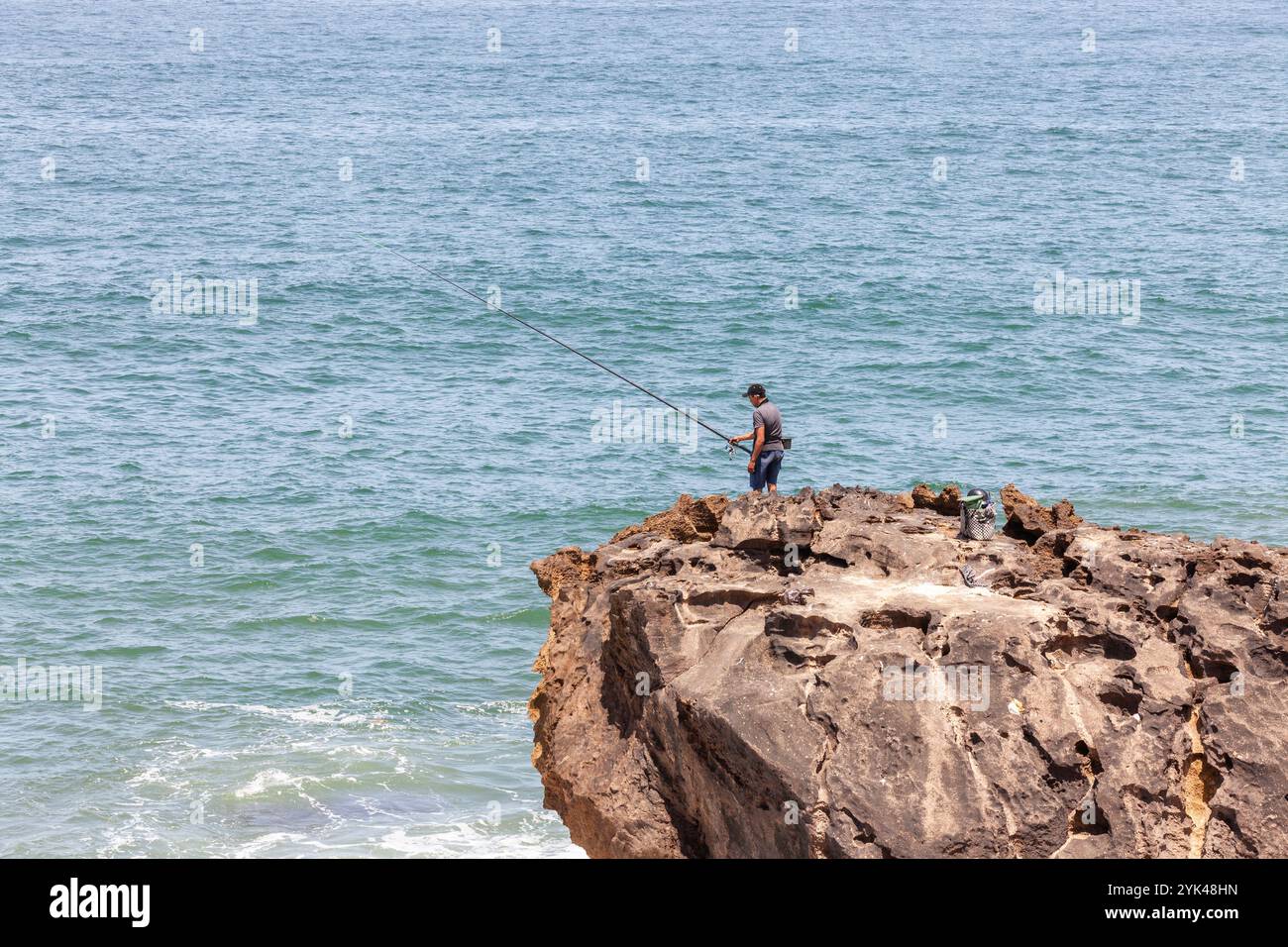 Ai margini di una scogliera rocciosa che si affaccia sul mare, un pescatore osserva la sua lenza da pesca Foto Stock